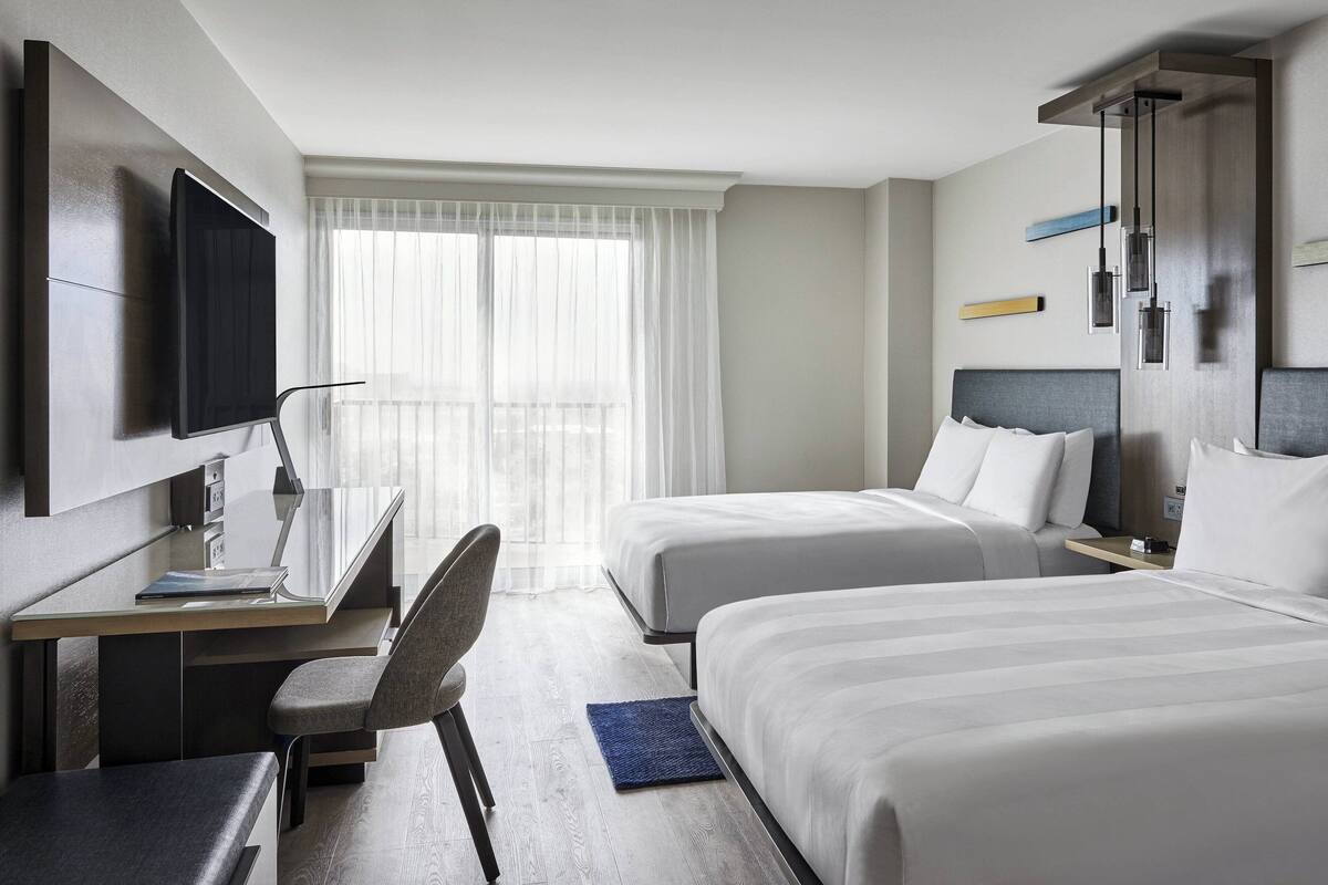 A hotel room with two beds, a desk, a chair, a TV, and a window with sheer curtains.