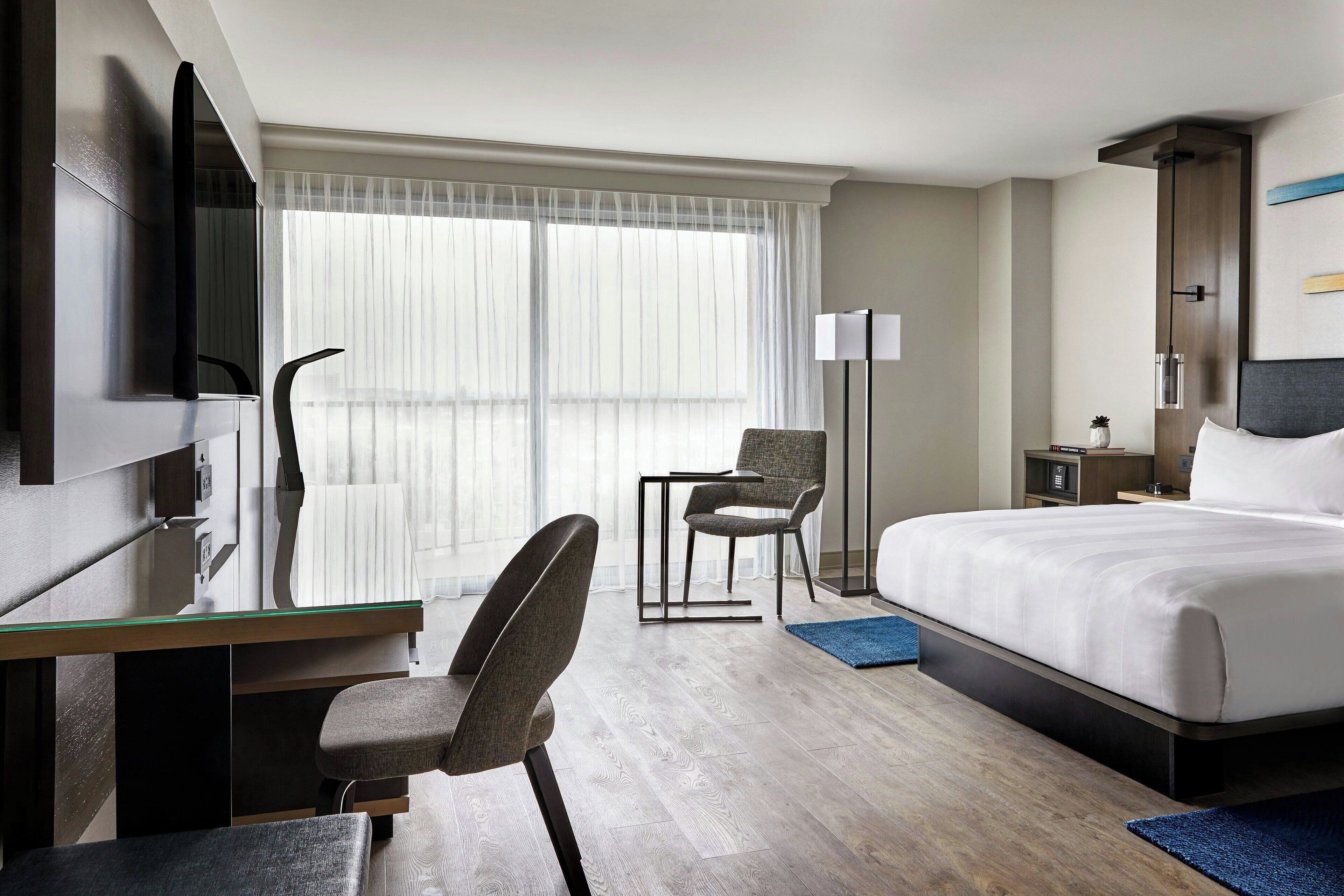 A hotel room with a bed, a desk with a chair, a TV, a lamp, and a window with sheer curtains.