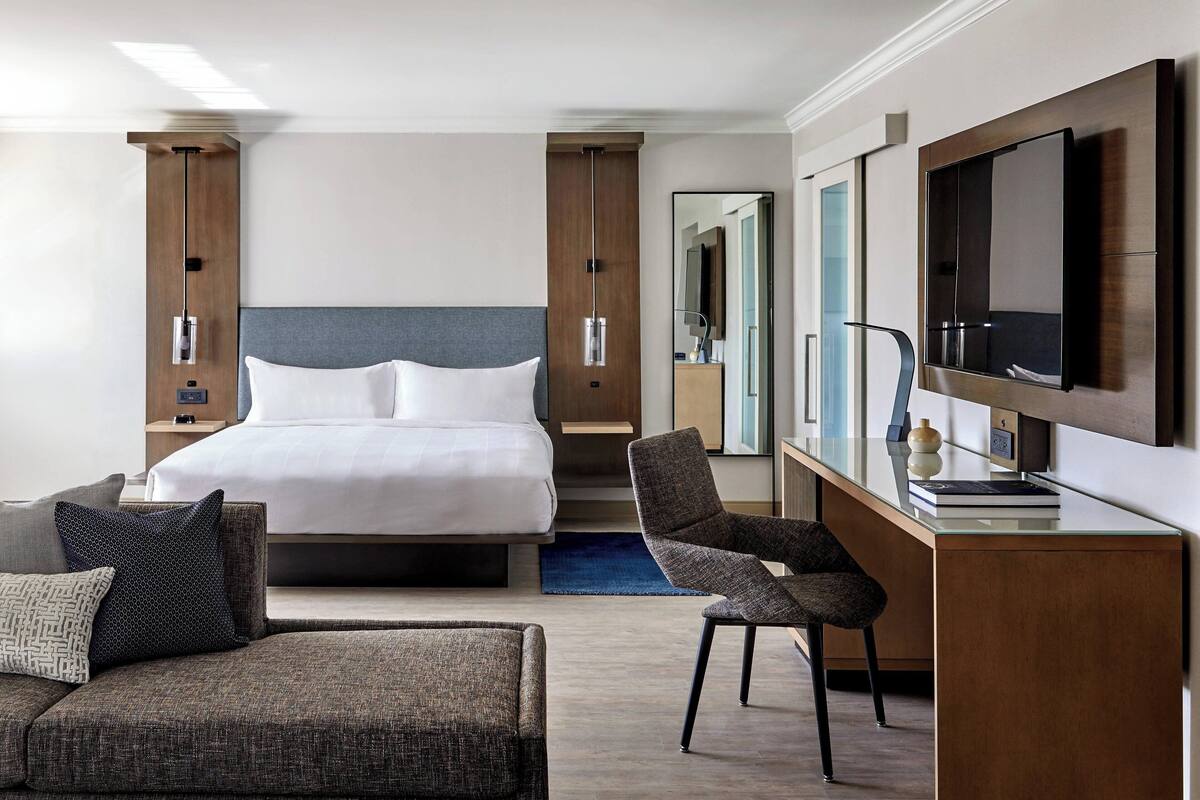A modern hotel room with a large bed, a desk with a flat-screen TV, a comfortable armchair, and a sofa.