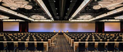 A conference room with rows of tables and chairs, a large screen at the front, and a ceiling with circular light fixtures.