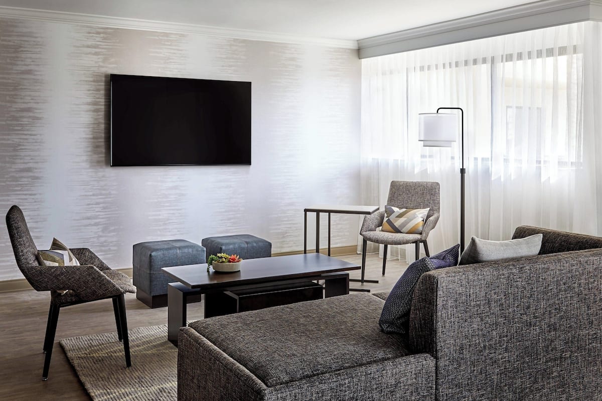 A modern living room with a flat-screen TV mounted on the wall, a sofa, a chair, a small table with a bowl of fruit, and a floor lamp.