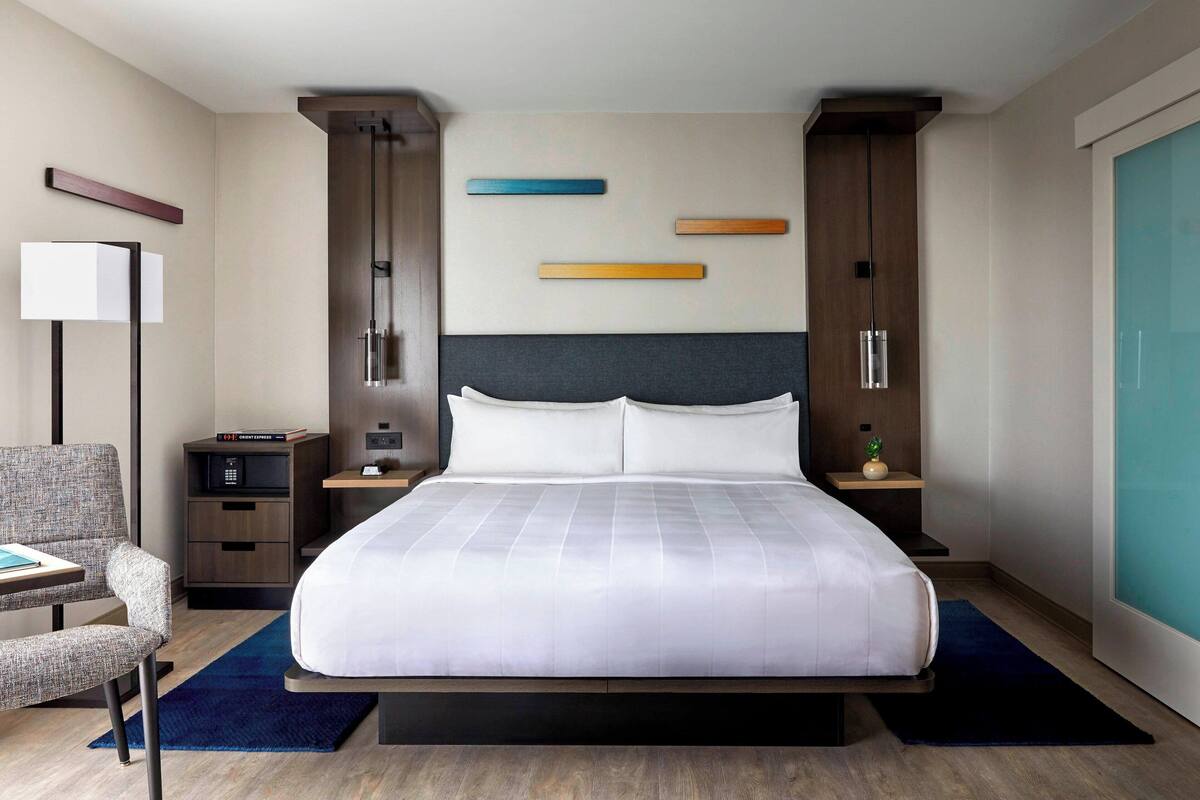 A modern hotel room with a large bed, a desk, a chair, a nightstand, a lamp, and a wall-mounted TV.