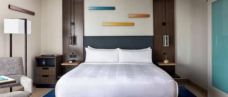 A modern hotel room with a large bed, a desk, a chair, a nightstand, a lamp, and a wall-mounted TV.