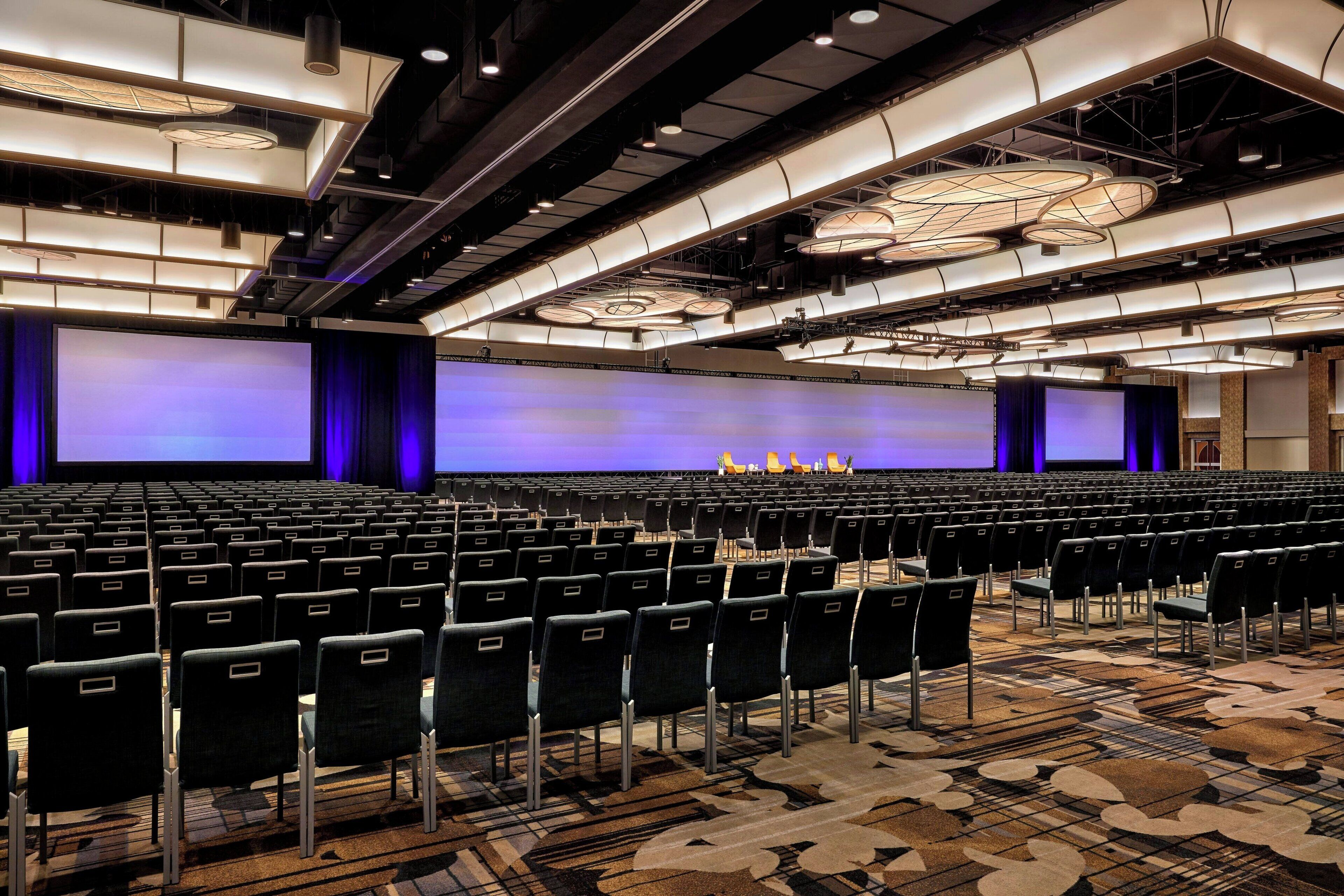 A large, modern conference room with rows of chairs, a large screen, and a stage with speakers.