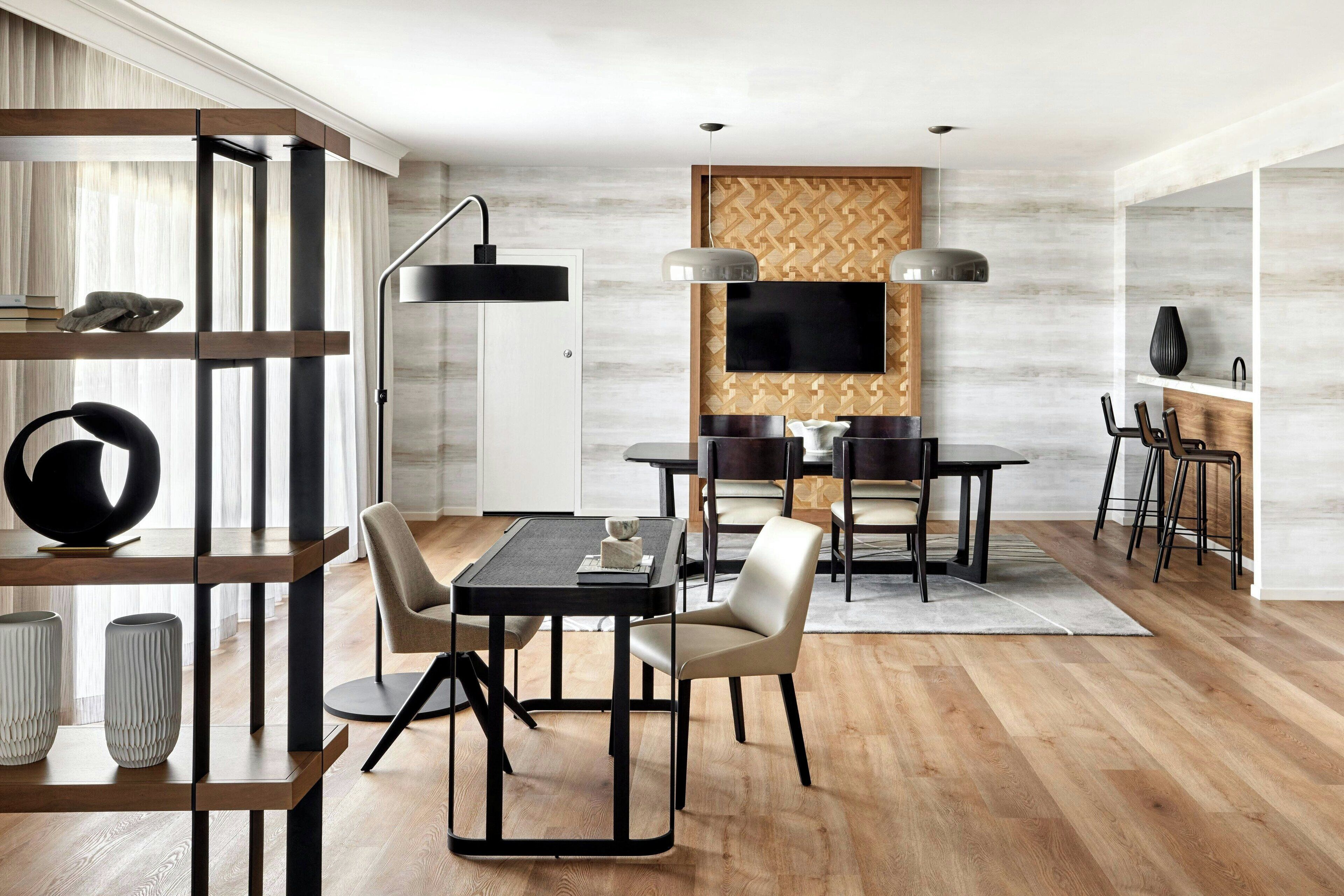 A modern dining area with a wooden floor, a black dining table, and chairs. There is a TV mounted on the wall and a bookshelf with decorative items.