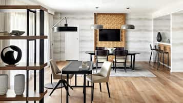 A modern dining area with a wooden floor, a black dining table, and chairs. There is a TV mounted on the wall and a bookshelf with decorative items.
