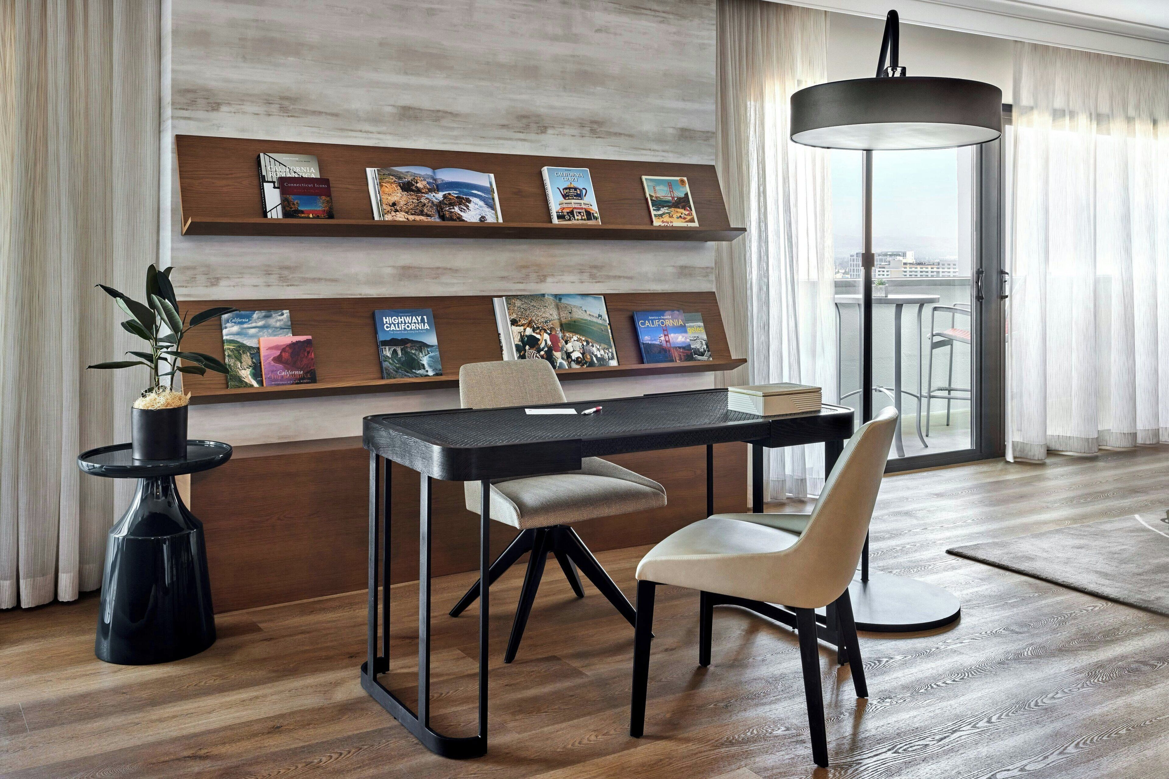 A modern dining area with a wooden table, two chairs, a potted plant, and a bookshelf with travel books.