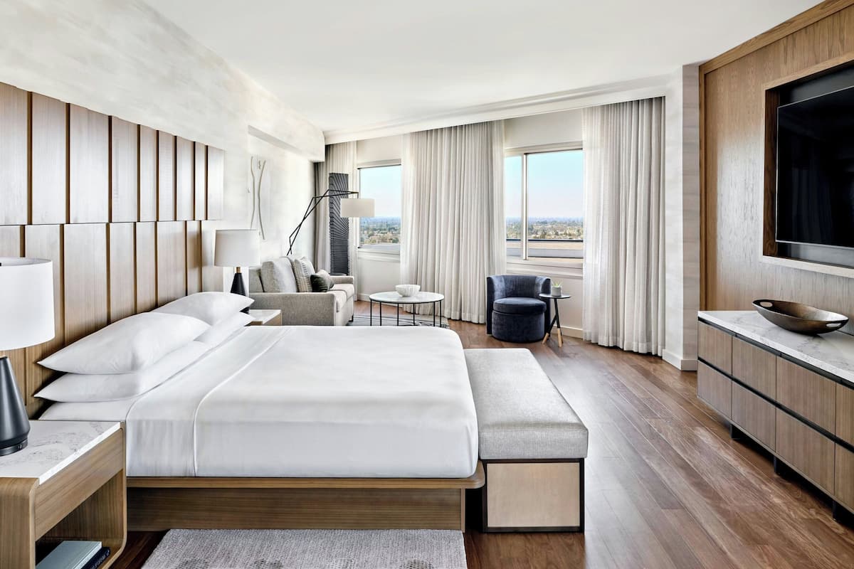 A modern hotel room with a large bed, a desk, a chair, a TV, and a view of the city.