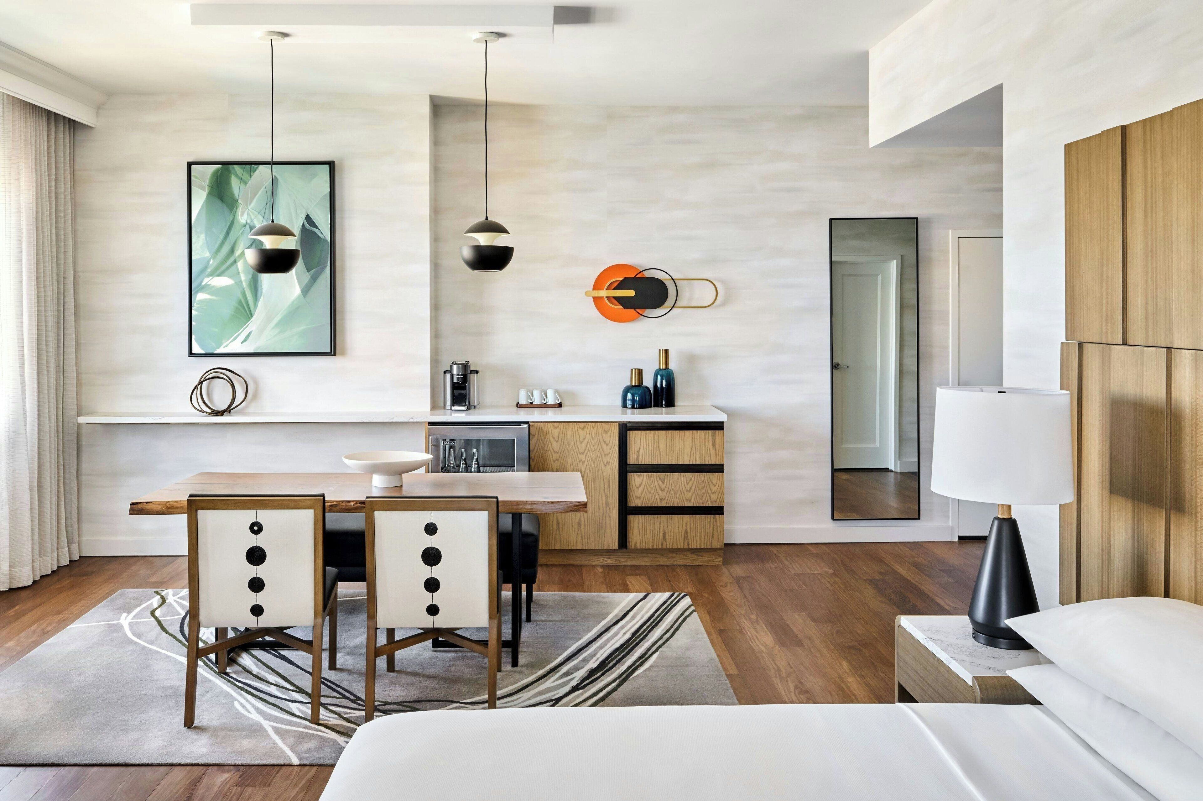 Une chambre d’hôtel moderne avec une table à manger en bois, deux chaises, un coin cuisine, un grand miroir et une lampe de chevet.