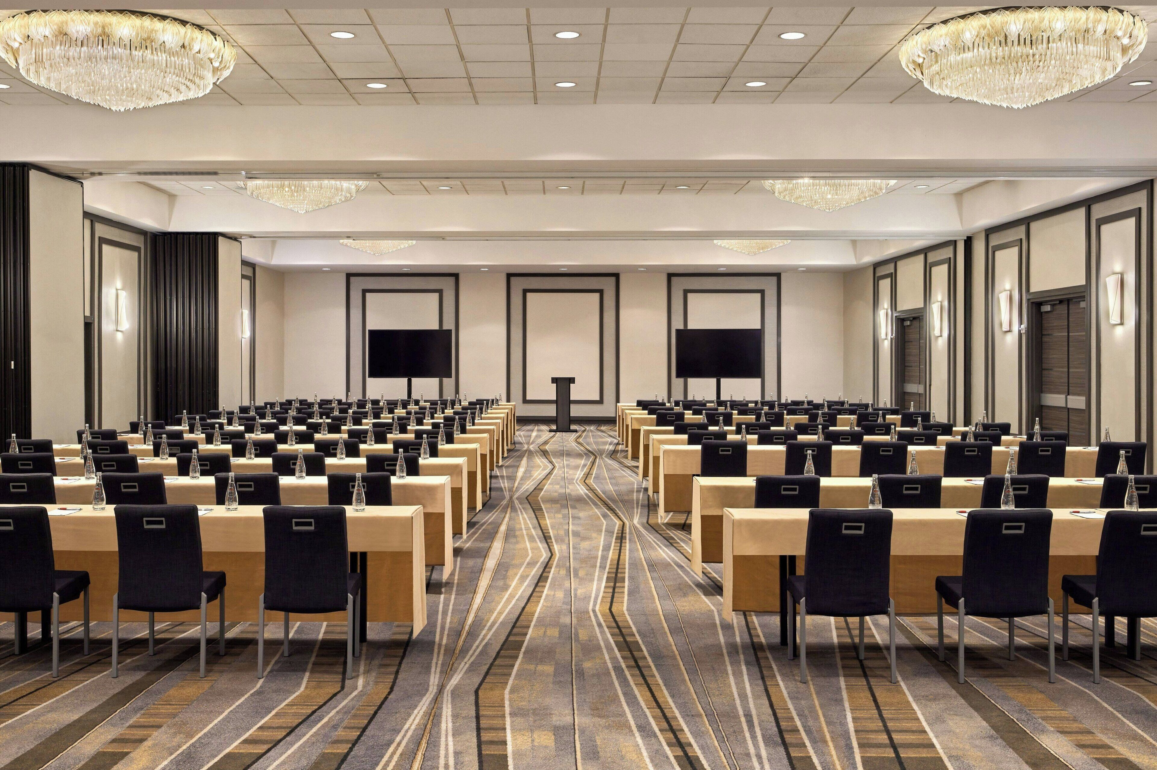 A conference room with rows of tables and chairs, large chandeliers, and a carpeted floor.