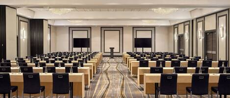 A conference room with rows of tables and chairs, large chandeliers, and a carpeted floor.