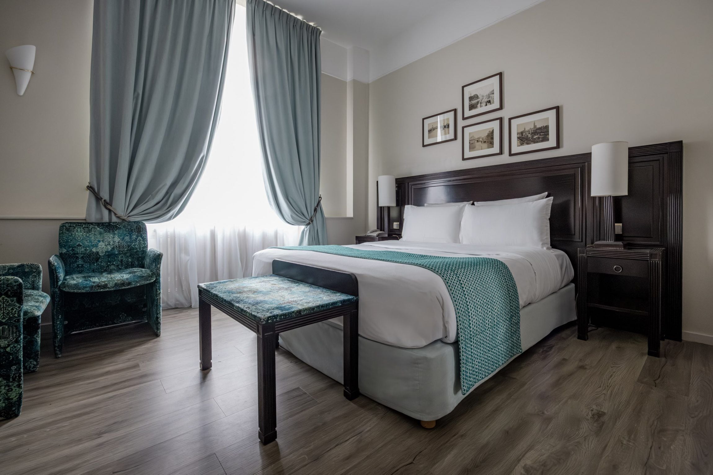 Una habitación de hotel con una cama grande, una silla, una mesita, una mesita de noche y un ventanal con cortinas.