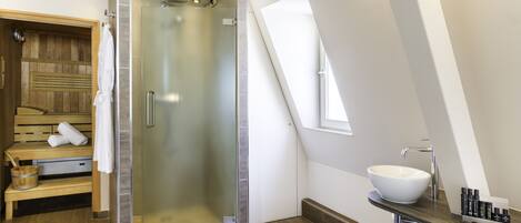 Uma casa de banho moderna com cabine de duche em vidro, banco de sauna em madeira, lavatório branco e toucador com produtos de higiene pessoal.