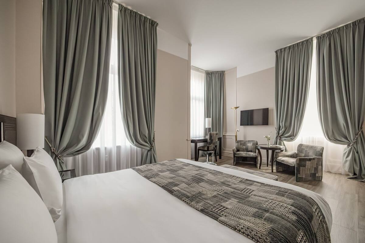 A hotel room with a large bed, two armchairs, a small table, a TV, and curtains.