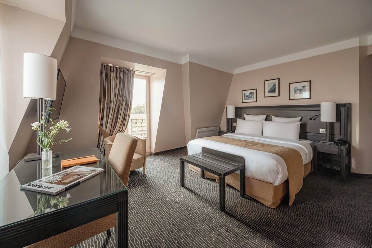 A hotel room with a bed, a desk, a chair, a small table, and a window with curtains.