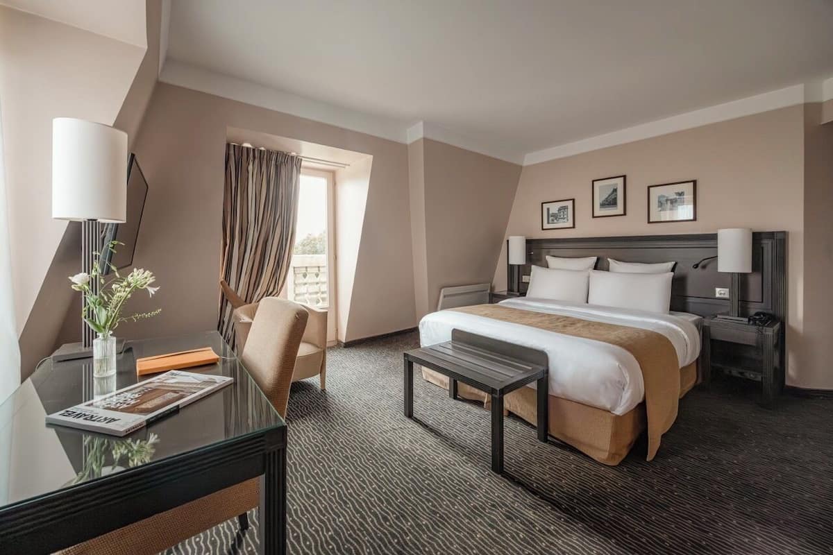 Quarto de hotel com uma cama, uma escrivaninha, uma cadeira, uma mesinha e uma janela com cortinas.