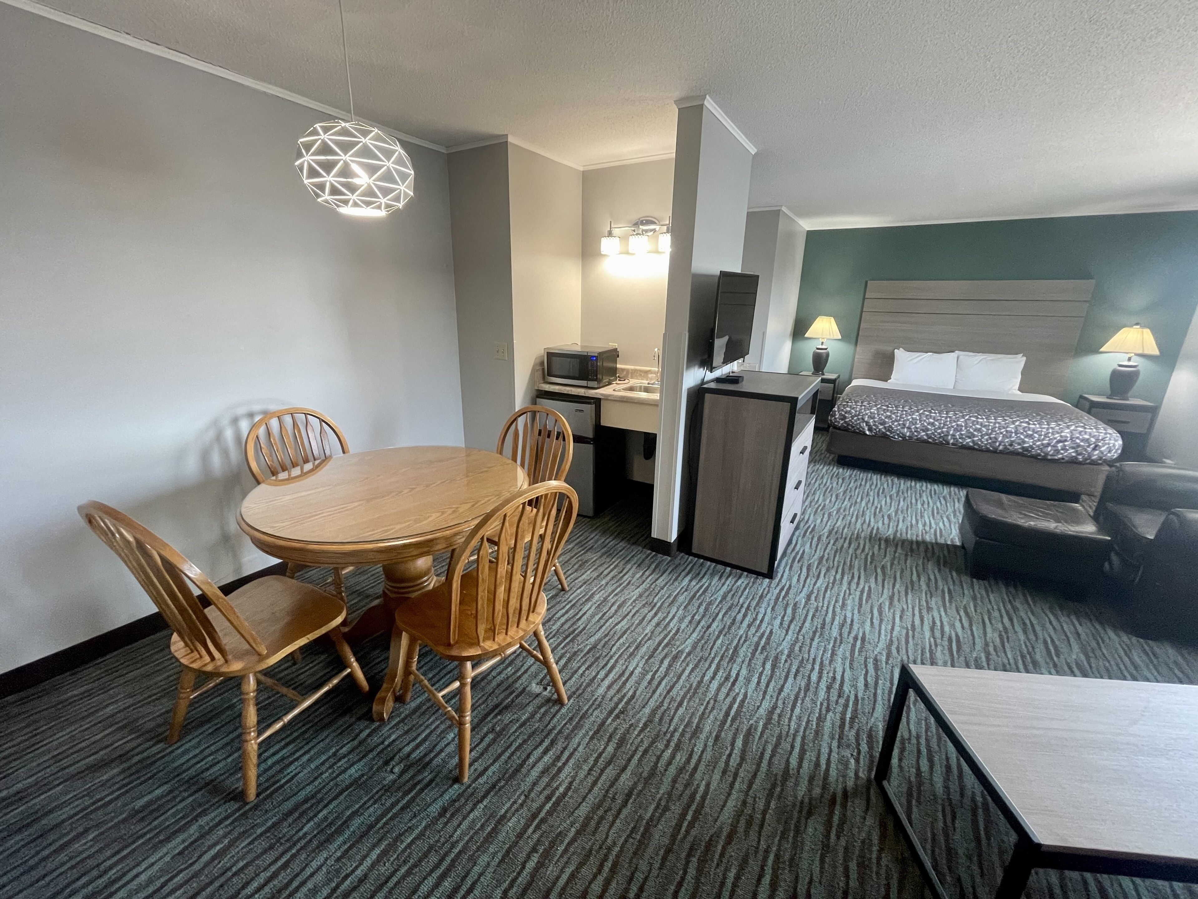 Habitación de hotel con mesa de comedor de madera, cuatro sillas, una cama con colcha estampada, un escritorio con computadora y microondas, y un mueble pequeño.