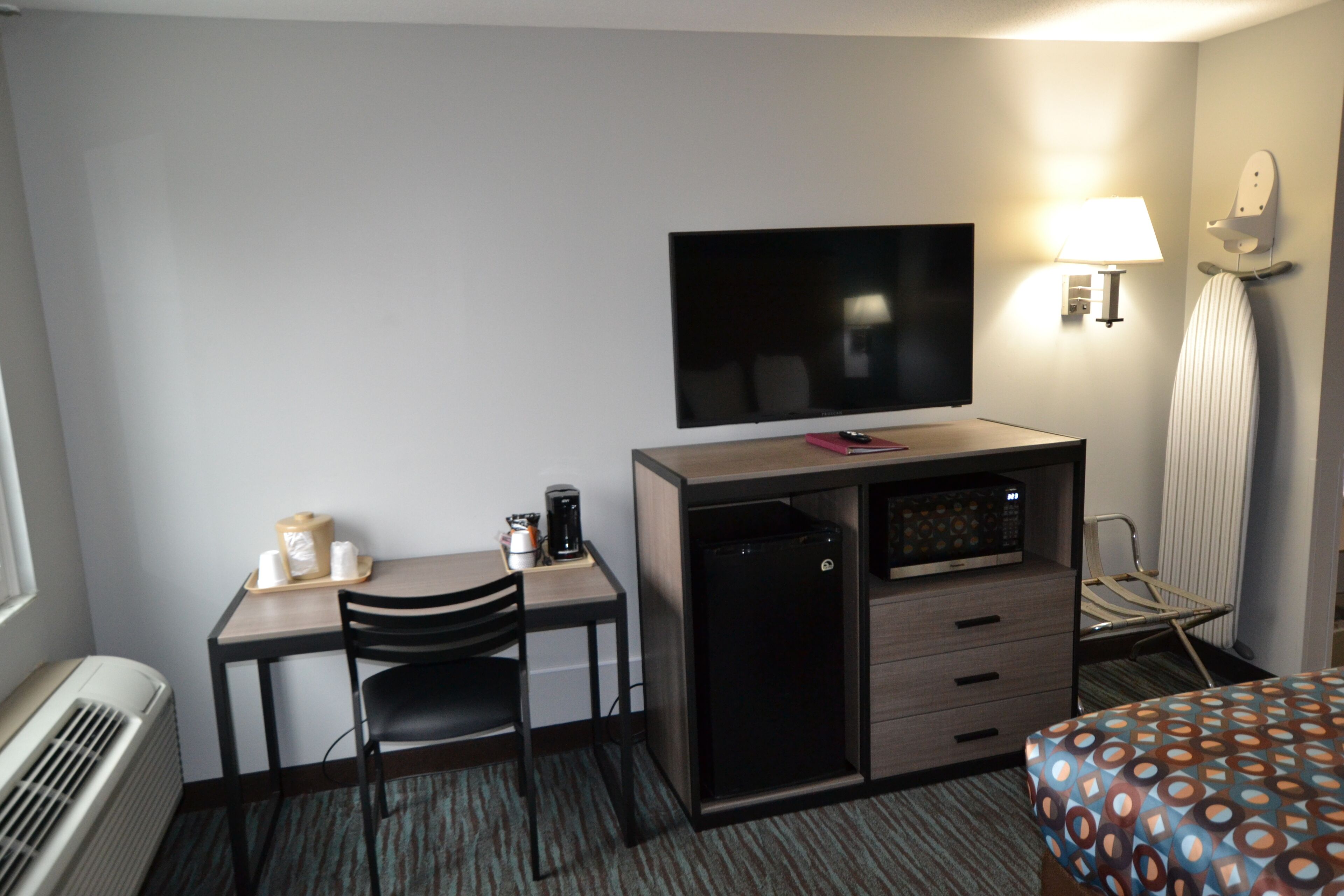 Habitación de hotel con escritorio, silla, televisión, mini refrigerador, lámpara, plancha y cama con edredón estampado.