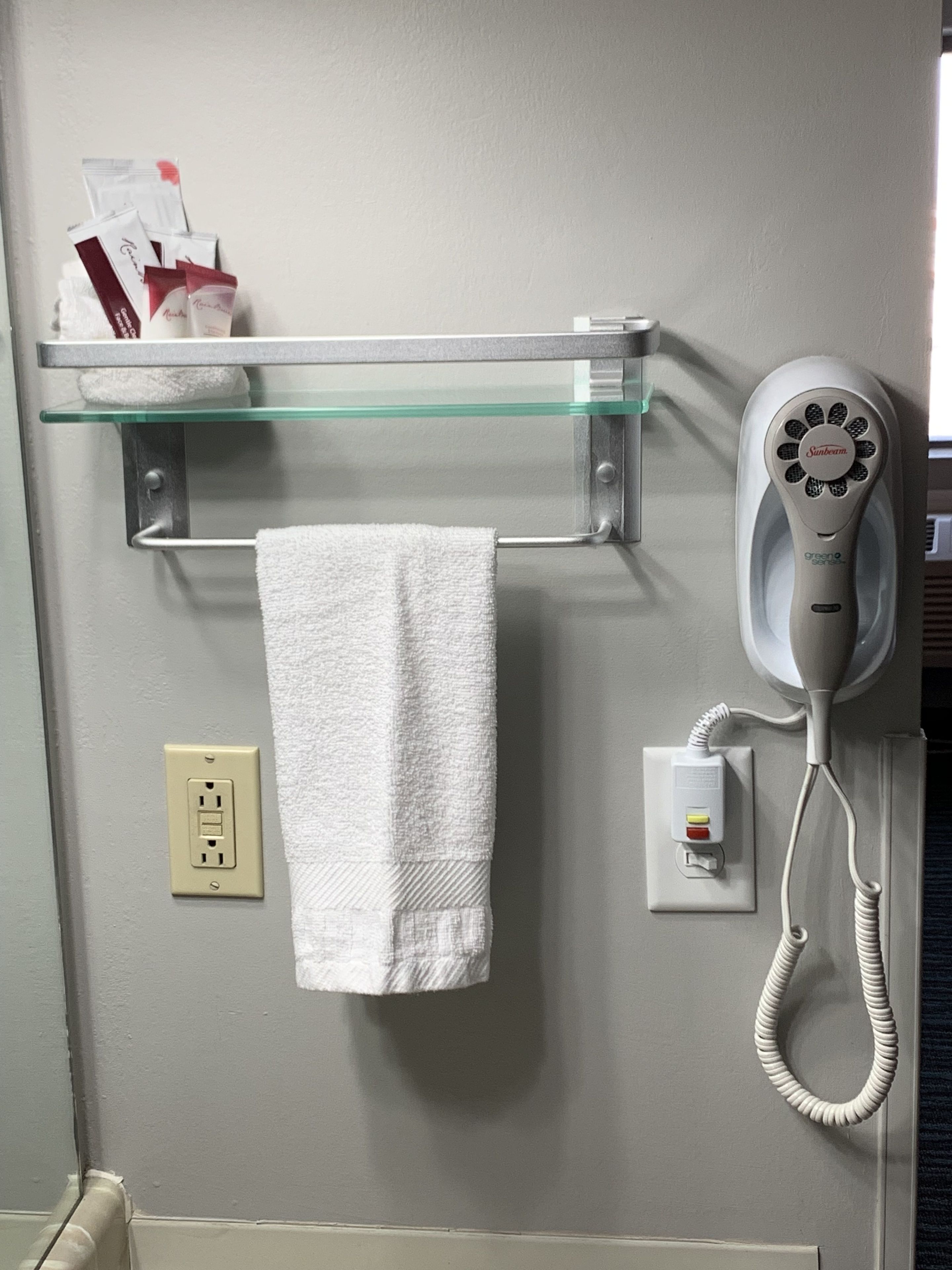 A bathroom with a glass shelf holding toiletries, a towel hanging on a rack, a hairdryer, and a power outlet.