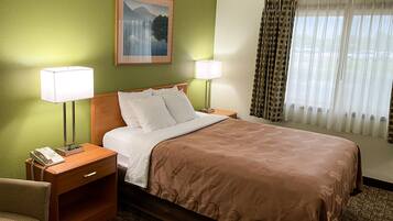 A hotel room with a bed, two bedside lamps, a telephone, a chair, a painting on the wall, and a window with curtains.
