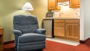 Un espacio habitable compacto con un sillón azul, un microondas, un refrigerador, armarios de madera y una lámpara.