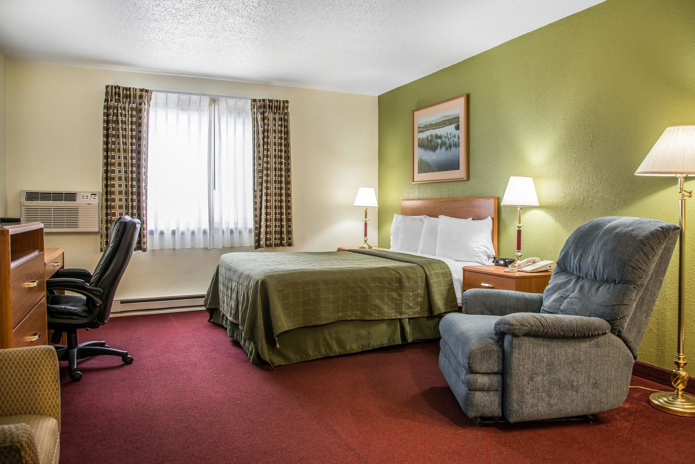 Una habitación de hotel con una cama, dos mesitas de noche, un escritorio, una silla y una ventana con cortinas.