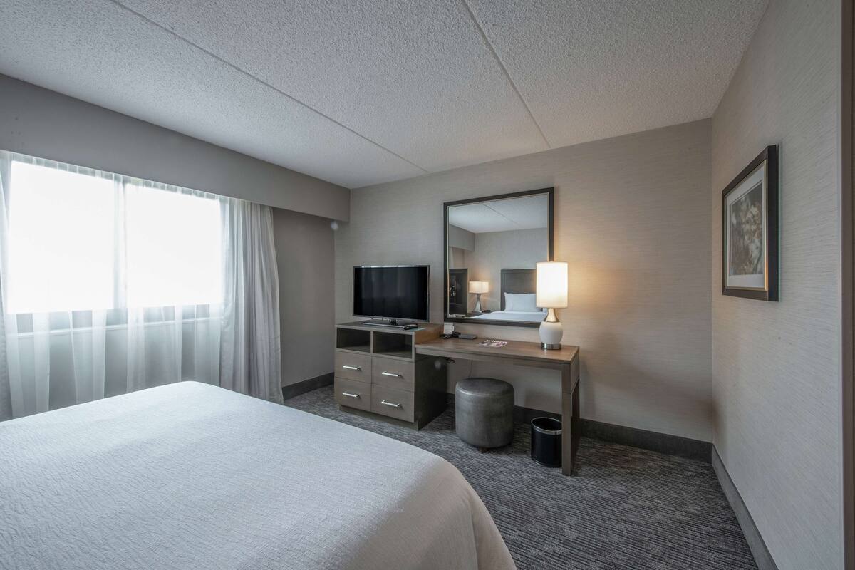 Habitación de hotel con cama, escritorio con televisor, lámpara, cubo de basura y un cuadro en la pared.