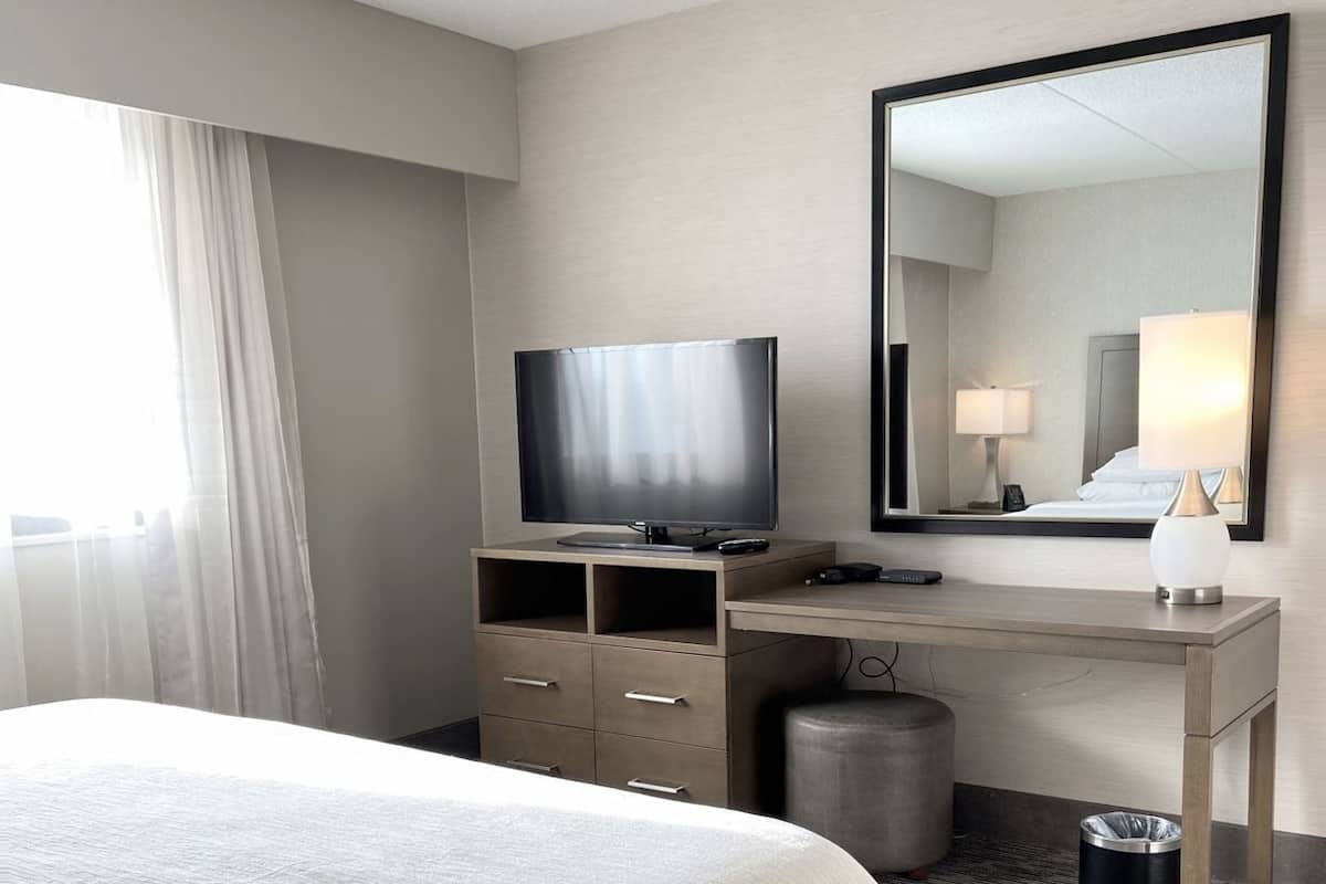 Habitación de hotel con cama, televisión, escritorio, espejo, lámpara, cubo de basura y ventana con cortinas.