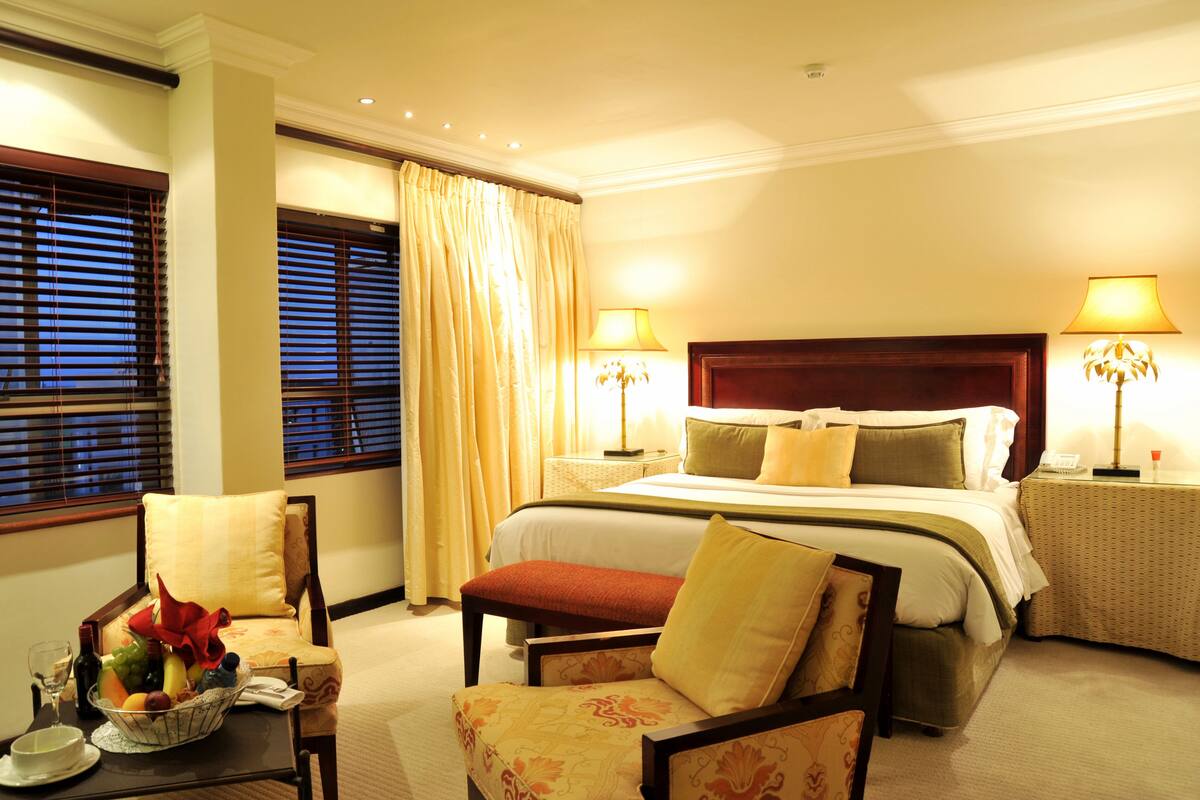 A hotel room with a large bed, two bedside lamps, a chair, a small table with a fruit bowl, and a window with blinds.