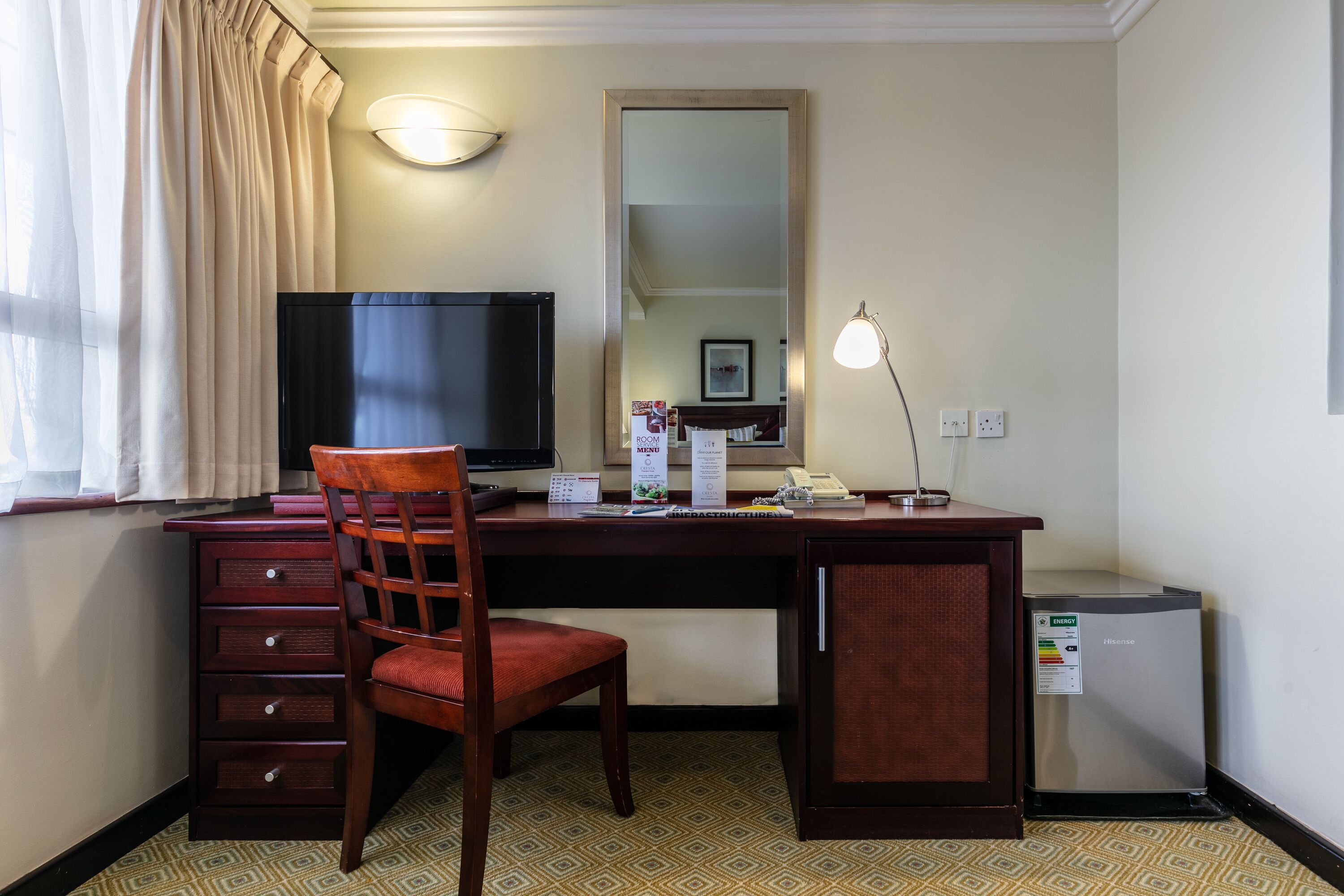 Habitación de hotel con un escritorio de madera, una silla, un televisor, un espejo, una lámpara y un mini refrigerador.