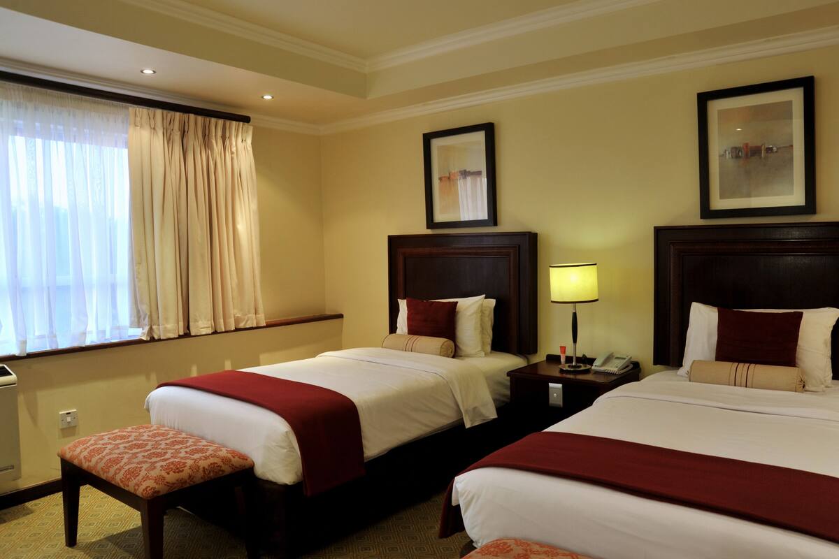 A hotel room with two beds, a nightstand with a lamp, a window with curtains, and a framed picture on the wall.