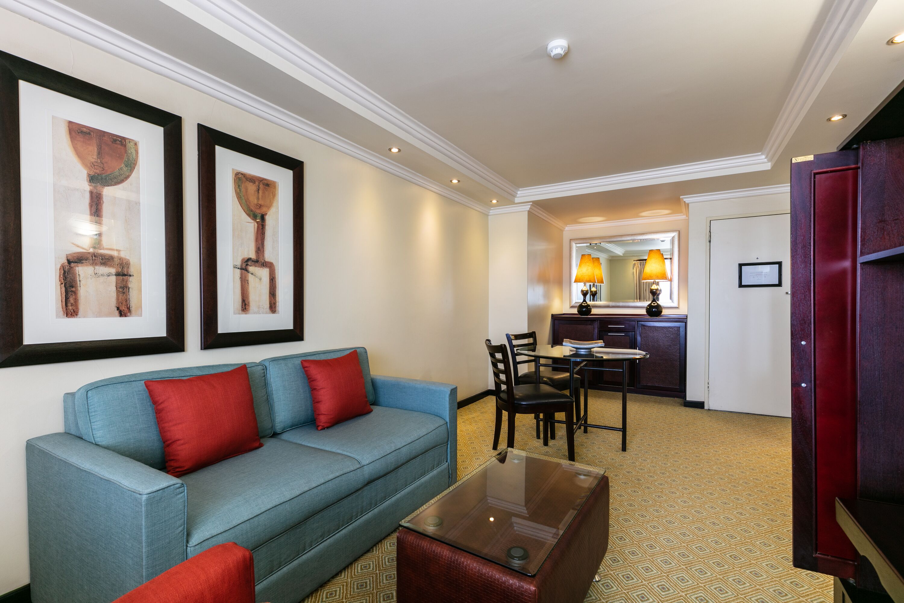 Habitación de hotel con un sofá azul, cojines rojos, una mesa de centro de vidrio y obras de arte enmarcadas en la pared.