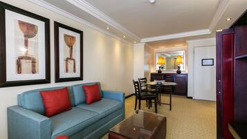 Habitación de hotel con un sofá azul, cojines rojos, una mesa de centro de vidrio y obras de arte enmarcadas en la pared.