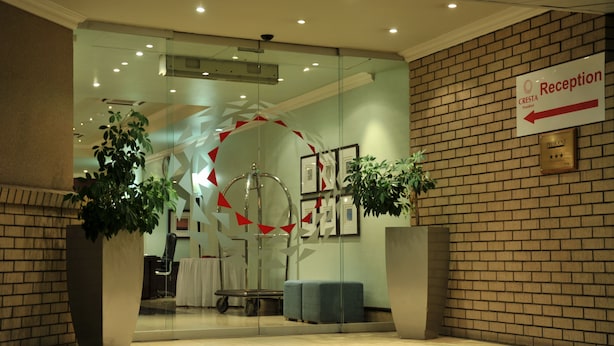 A reception area with a brick wall, a reception desk, and two large potted plants. - Cresta President (Gaborone)