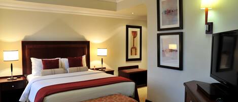 Una habitación de hotel con una cama grande, dos mesitas de noche con lámparas, un banco, un televisor y cuadros en las paredes.