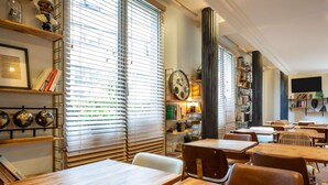 A restaurant interior with wooden tables and chairs, large windows with blinds, and shelves with books and decorative items. - ibis Styles Paris Tolbiac Bibliotheque (Paris)