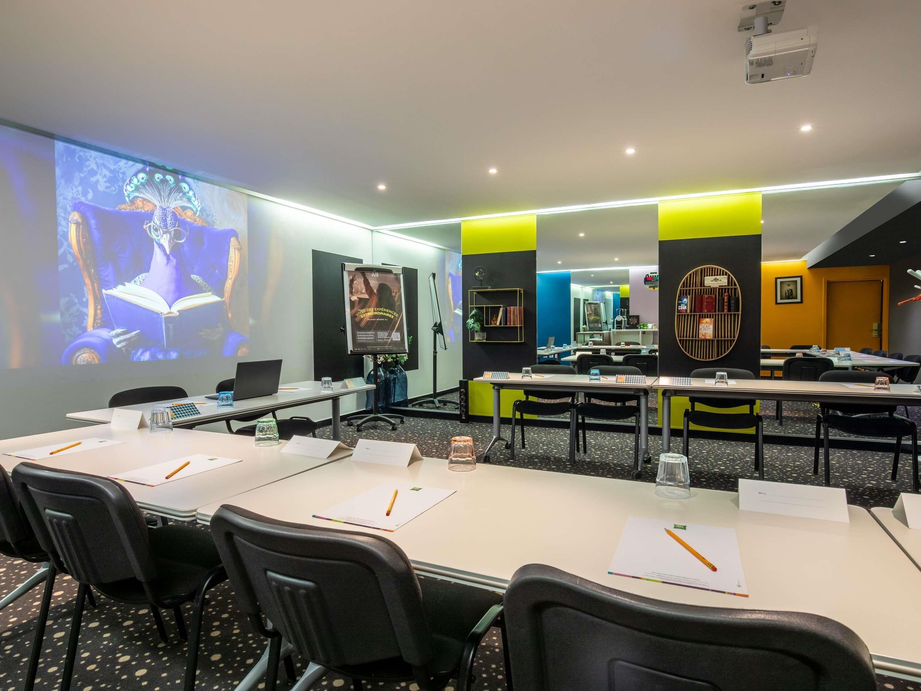 A conference room with tables, chairs, a projector screen, and a large mirror.