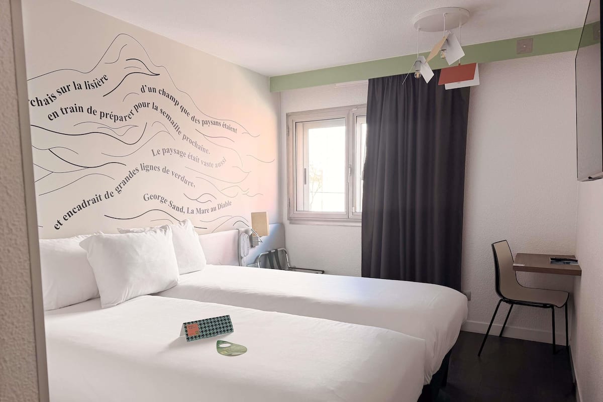 Ein Hotelzimmer mit zwei Betten, einem Schreibtisch, einem Sessel, einem Fenster mit Vorhängen und einer Wand mit einer dekorativen Zitat.