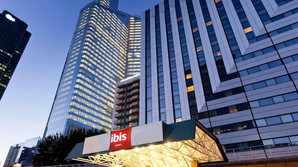 A modern hotel with a prominent glass facade, a distinctive geometric entrance, and a visible signage for 'ibis Hotels'. - ibis Paris La Défense Esplanade (Courbevoie)