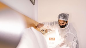 A person in a white robe and hairnet attending to a bed in a room with a sign on the wall. - ibis Paris La Défense Esplanade (Courbevoie)