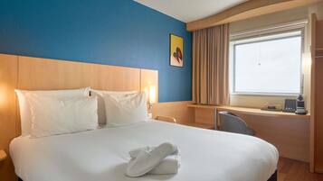 A hotel room with a large bed, a desk, a chair, a window with curtains, and a painting on the wall.