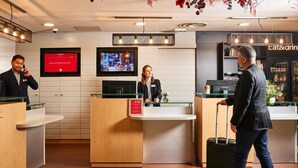 A hotel reception area with a reception desk, a TV screen, and a bar area in the background. - ibis Paris La Défense Esplanade (Courbevoie)