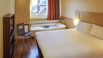 Habitación de hotel con cama, una silla, escritorio, ventana con cortinas y vista a edificios.