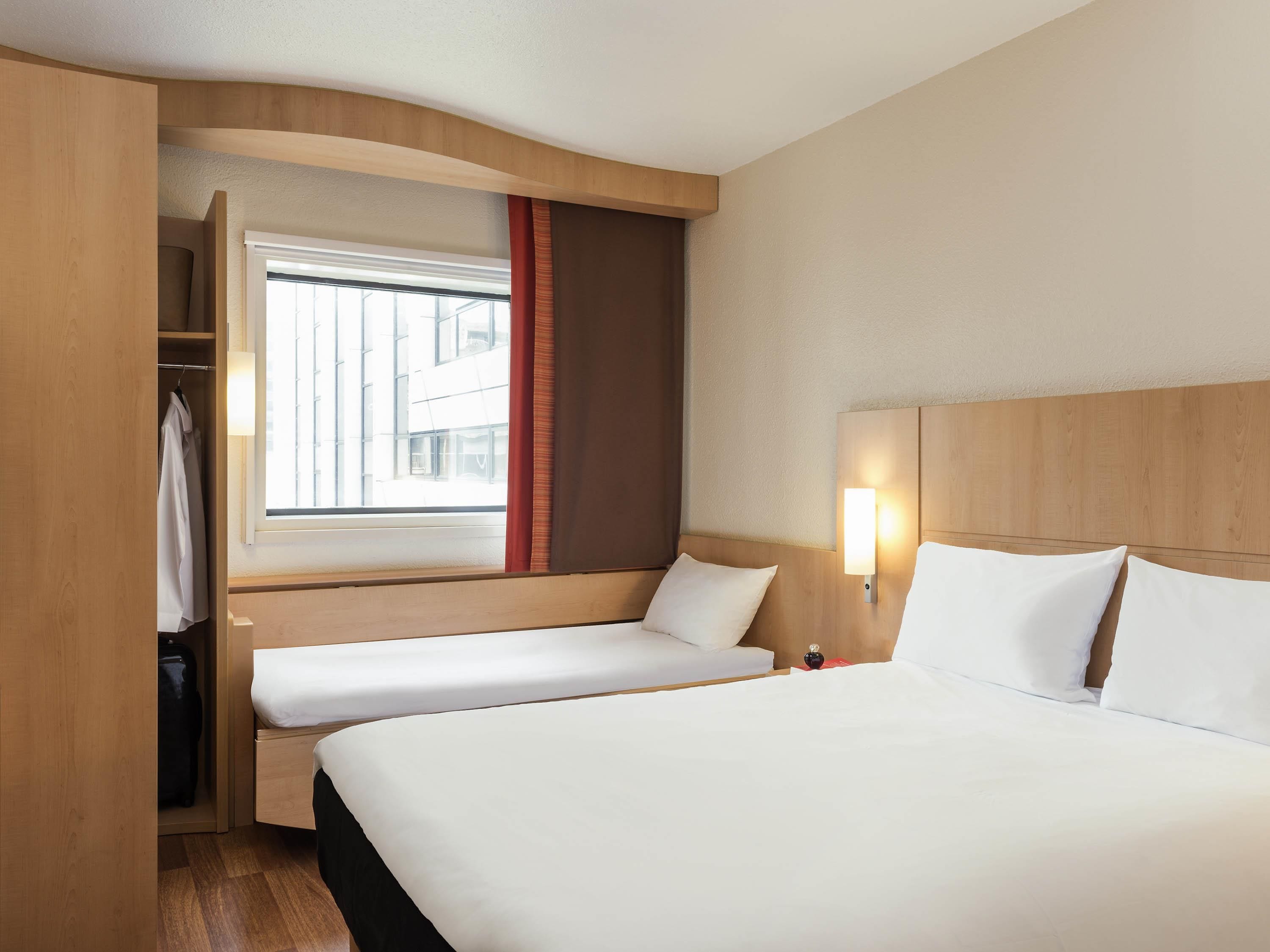 A hotel room with a large bed, a smaller bed, a wardrobe, a window with curtains, and a wall-mounted light.