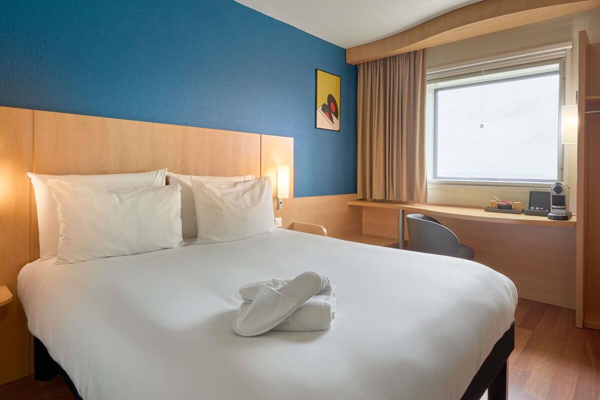 Une chambre d’hôtel avec un grand lit, un bureau, une chaise, une fenêtre avec des rideaux et un tableau au mur.