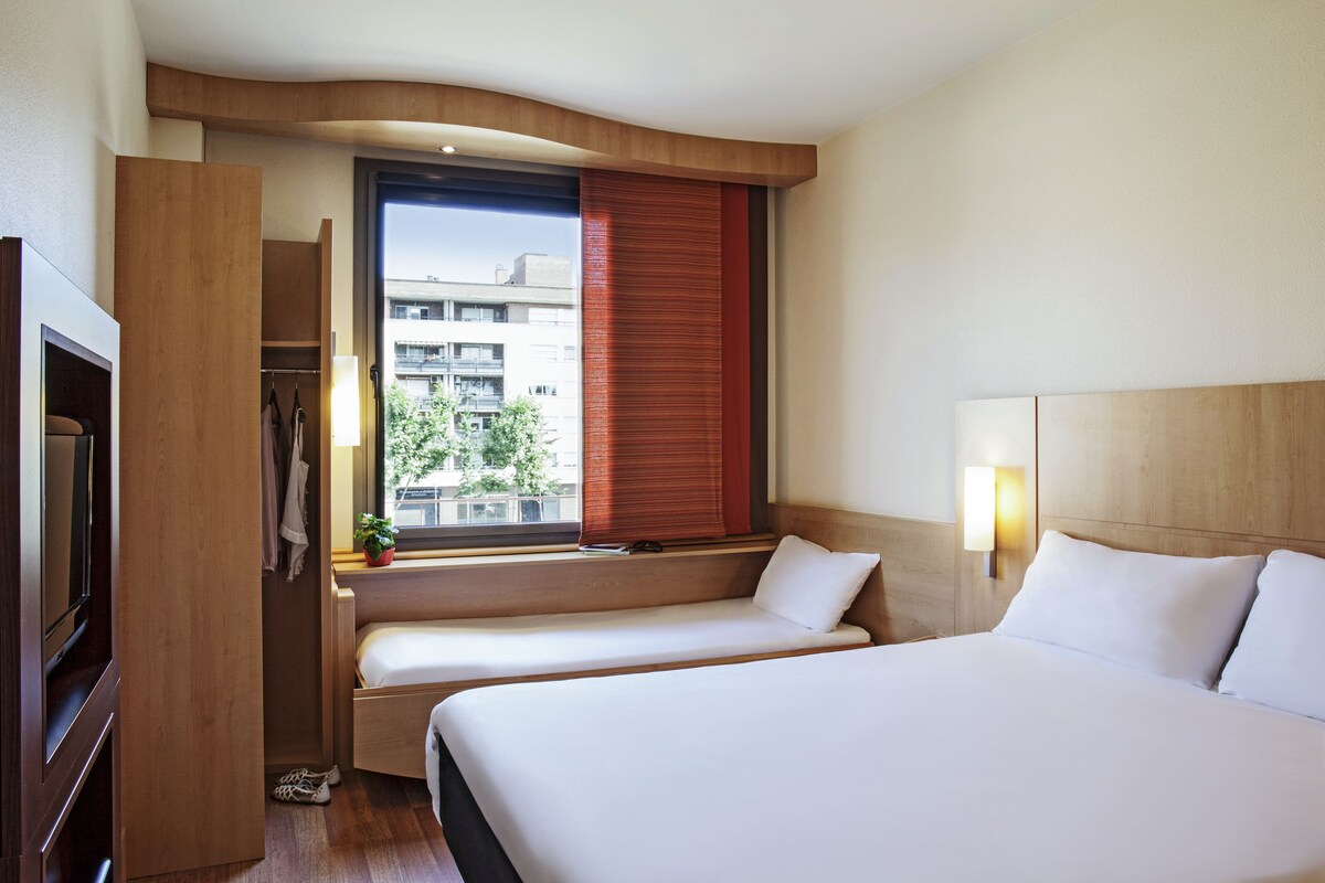 A hotel room with two beds, a wardrobe, a window with a view of buildings, and a bedside lamp.