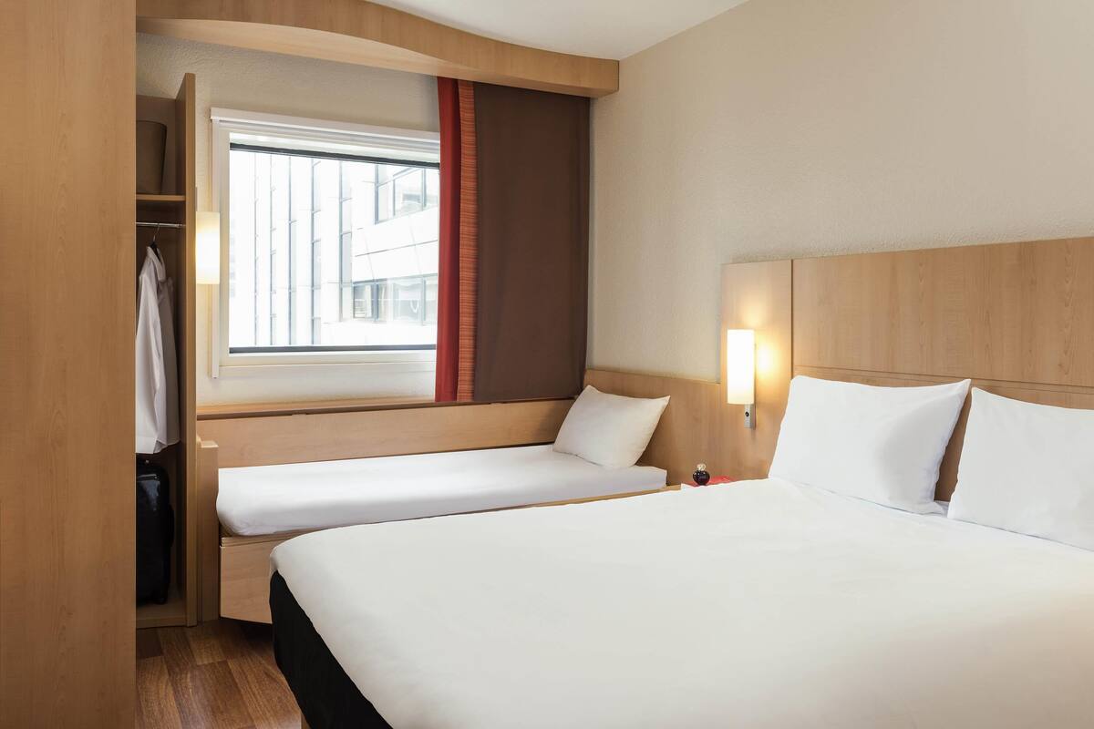 Une chambre d’hôtel avec un grand lit, un lit plus petit, une armoire, une fenêtre avec des rideaux et un luminaire fixé au mur.
