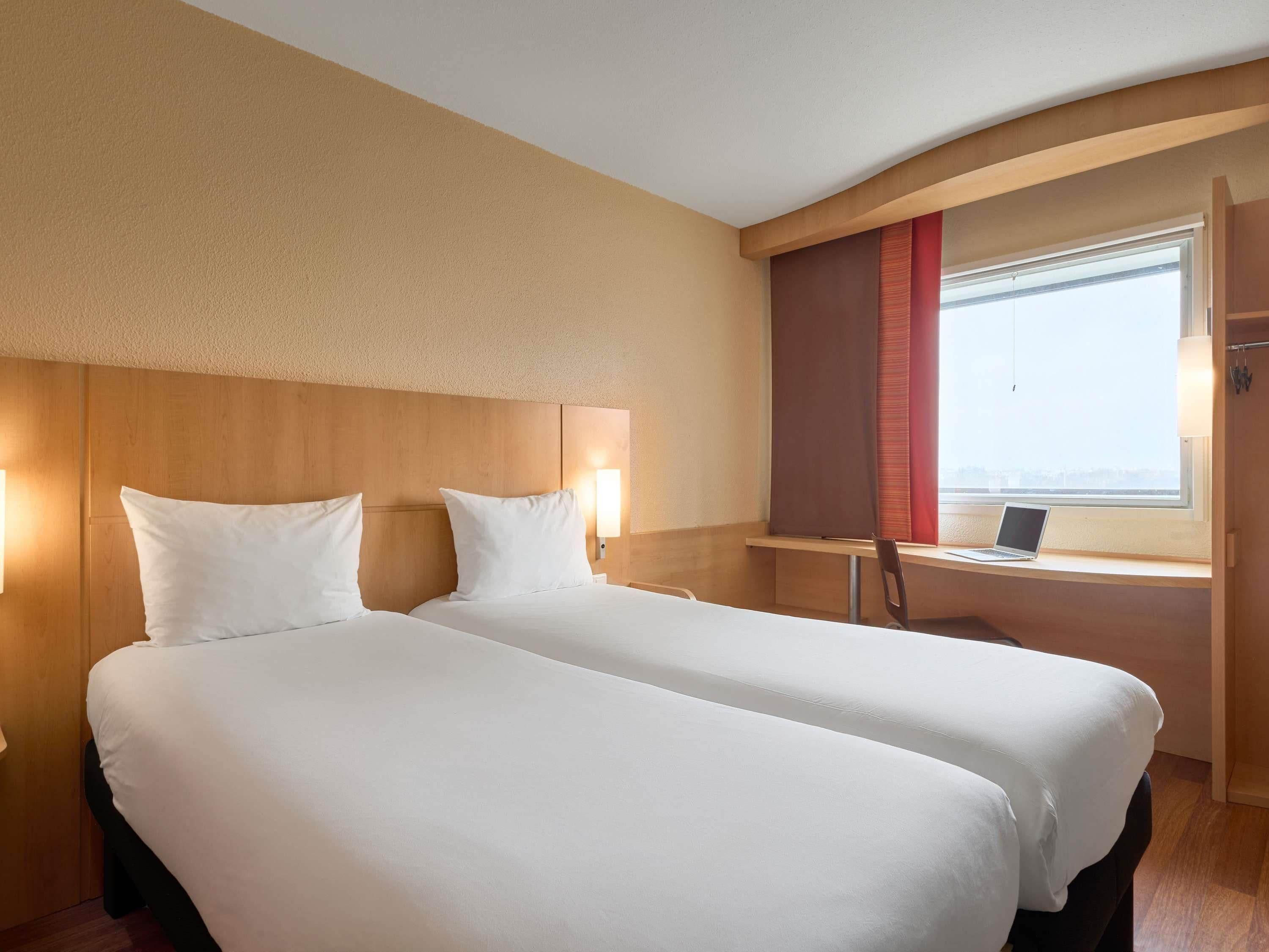 A hotel room with two beds, a desk, a laptop, and a window with a view.