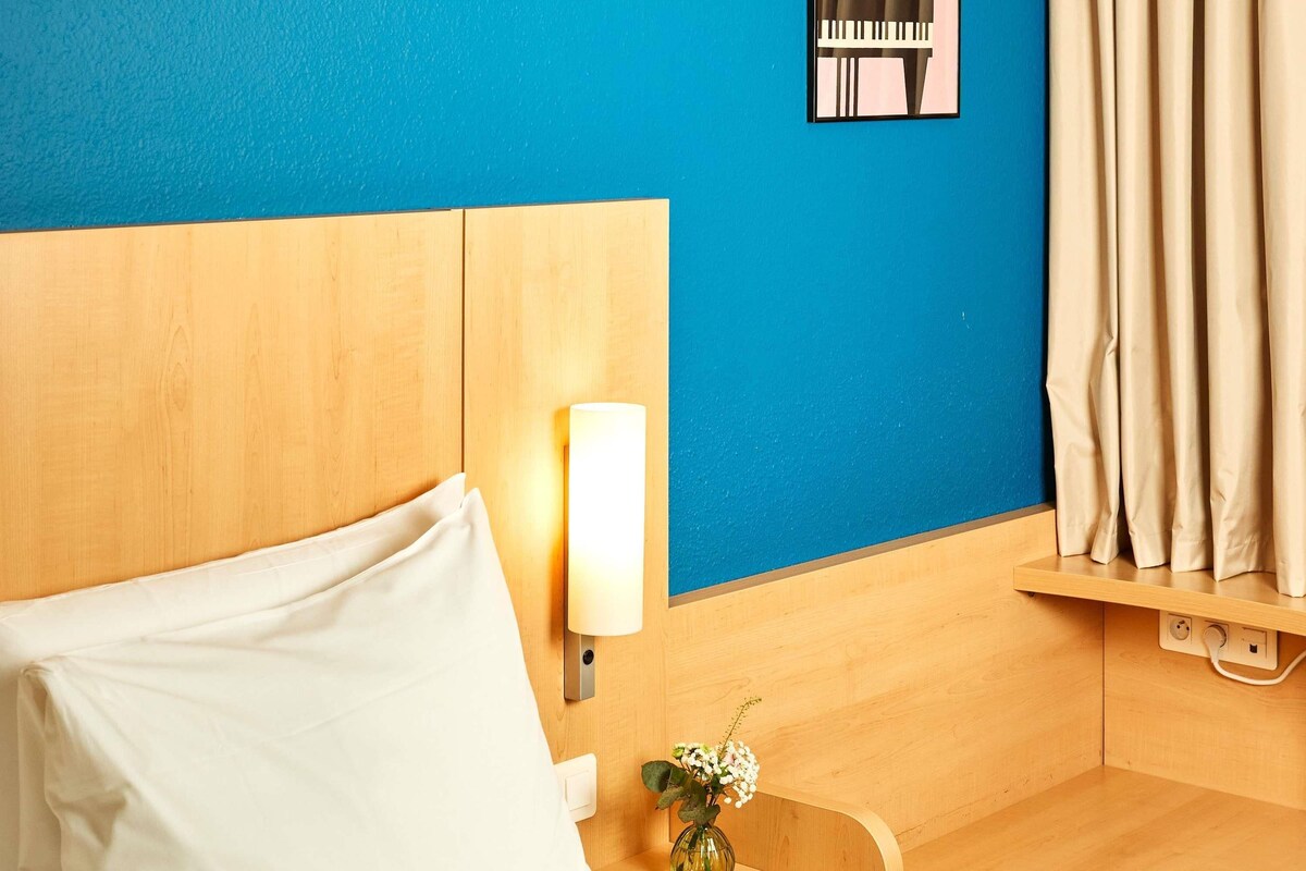 Quarto de hotel com cabeceira de madeira, luminária de cabeceira, mesinha com um vaso de flores e cortina na janela.