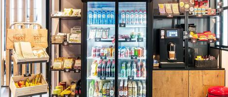 A beverage refrigerator with a sign above it, a display of snacks and beverages, and a coffee station with a red trash bin.